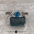 The "Saorise" ring in 18K white gold, set with a 0.90-carat Montana sapphire and Celtic knot design, rests on a rough blue-green stone atop a textured gray surface. The "Earth's Treasury" logo appears at the bottom.