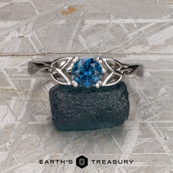 The "Saorise" ring in 18K white gold with 0.90-carat Montana sapphire The "Saorise" ring in 18K white gold, set with a 0.90-carat Montana sapphire and Celtic knot design, rests on a rough blue-green stone atop a textured gray surface. The "Earth's Treasury" logo appears at the bottom.