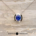 A 14K white gold "The Cirrus" Necklace featuring a 2.73-carat cushion-cut tanzanite, accented by small clear stones, displayed on light wood with the "Earth's Treasury" logo at the bottom. Build your own unique pendant style today.