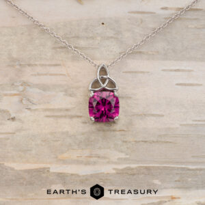 The "Saoirse" pendant in 14K white gold features a Celtic knot design and a 1.95-carat Mahenge garnet, shown against light wood with the Earth's Treasury logo. Ideal for anyone looking for unique, custom jewelry.