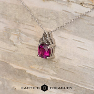 The "Saoirse" pendant in 14K white gold features a Celtic knot design and a 1.95-carat Mahenge garnet, shown on light wood with the "Earth's Treasury" logo—ideal for custom jewelry enthusiasts.