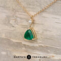 The "Raina" Necklace in 14K yellow gold with a 1.46-carat emerald and a small clear stone, displayed on light wood with The Earth's Treasury logo—ideal for custom jewelry lovers.