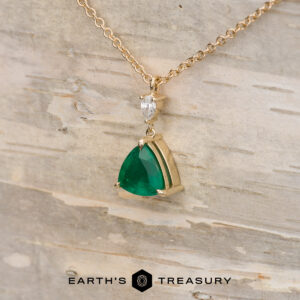 The "Raina" Necklace in 14K yellow gold with a 1.46-carat emerald and a small clear stone, displayed on light wood with The Earth's Treasury logo—ideal for custom jewelry lovers.
