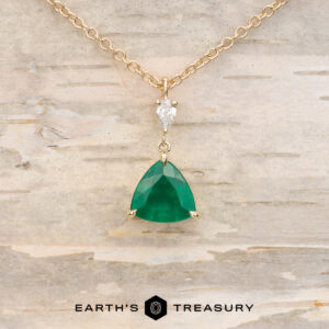 The "Raina" Necklace in 14K yellow gold features a pear-shaped white gemstone above a 1.46-carat triangular emerald pendant, displayed on a light textured surface with "EARTH'S TREASURY" below. Build your own custom jewelry with this elegant piece.