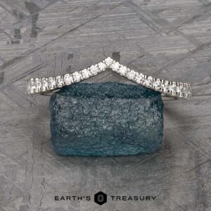 The Chevron Tapered Pave Diamond Band in platinum stands on a rough blue gemstone, set against a geometric background with the "Earth's Treasury" logo below.