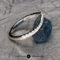 The Chevron Tapered Pave Diamond Band in platinum, featuring a row of small diamonds, is displayed leaning against a rough blue gemstone on a textured gray surface. "Earth's Treasury" appears at the bottom.