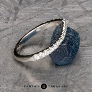 The Chevron Tapered Pave Diamond Band in platinum, featuring a row of small diamonds, is displayed leaning against a rough blue gemstone on a textured gray surface. "Earth's Treasury" appears at the bottom.
