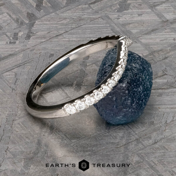 The Chevron Tapered Pave Diamond Band in platinum The Chevron Tapered Pave Diamond Band in platinum, featuring a row of small diamonds, is displayed leaning against a rough blue gemstone on a textured gray surface. "Earth's Treasury" appears at the bottom.