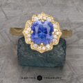 The "Mirabelle" ring in 18K yellow gold features a 1.92-carat Ceylon sapphire at the center, encircled by sparkling white diamonds in a floral halo, shown on a textured dark stone background.