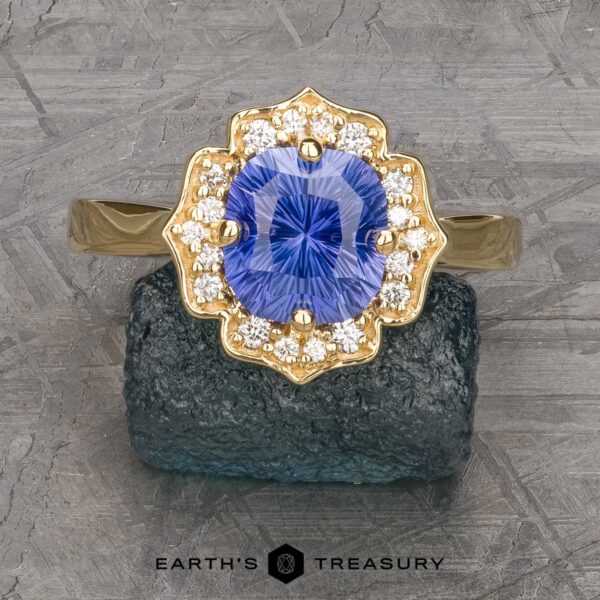 The "Mirabelle" ring in 18K yellow gold with 1.92-Carat Ceylon sapphire The "Mirabelle" ring in 18K yellow gold features a 1.92-carat Ceylon sapphire at the center, encircled by sparkling white diamonds in a floral halo, shown on a textured dark stone background.
