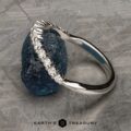 The Tapered Prong-Set Diamond Contour Band in 14K white gold, featuring a row of sparkling diamonds, is shown resting on a rough blue stone with a textured gray background and the Earth’s Treasury logo below.
