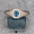 The "Gawain" ring in unplated 18k palladium white gold, set with a 0.79-carat Montana sapphire, stands upright on a rough blue stone. Its geometric textured design is shown against a gray patterned background.