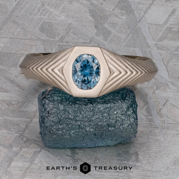The "Gawain" ring in unplated 18k palladium white gold with 0.79-Carat Montana sapphire The "Gawain" ring in unplated 18k palladium white gold, set with a 0.79-carat Montana sapphire, stands upright on a rough blue stone. Its geometric textured design is shown against a gray patterned background.