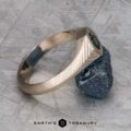 The "Gawain" ring in unplated 18k palladium white gold, featuring a 0.79-carat Montana sapphire, is shown leaning against a rough blue stone on a textured gray surface with the Earth’s Treasury logo at the bottom.