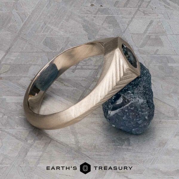 The "Gawain" ring in unplated 18k palladium white gold with 0.79-Carat Montana sapphire The "Gawain" ring in unplated 18k palladium white gold, featuring a 0.79-carat Montana sapphire, is shown leaning against a rough blue stone on a textured gray surface with the Earth’s Treasury logo at the bottom.