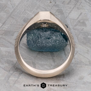 The "Gawain" ring, crafted in unplated 18k palladium white gold with a 0.79-carat Montana sapphire, is shown upright on a textured gray surface. The Earth's Treasury logo appears at the bottom.