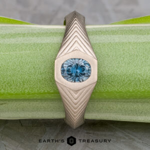 The "Gawain" ring, crafted in unplated 18k palladium white gold with a 0.79-carat Montana sapphire, features geometric patterns along the band and is displayed on a green textured surface.