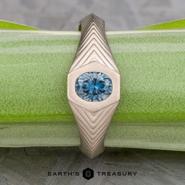 The "Gawain" ring in unplated 18k palladium white gold with 0.79-Carat Montana sapphire The "Gawain" ring, crafted in unplated 18k palladium white gold with a 0.79-carat Montana sapphire, features geometric patterns along the band and is displayed on a green textured surface.