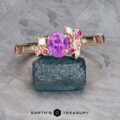 The Linnea ring in 14K yellow gold features a 1.15-carat Ceylon sapphire center, surrounded by pink and white accent stones, displayed on a rough dark blue crystal with the "EARTH'S TREASURY" logo below.