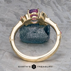 The Linnea ring in 14K yellow gold, featuring a 1.15-carat Ceylon sapphire and clear side stones, stands upright on a textured metallic surface with the Earth's Treasury logo displayed below.