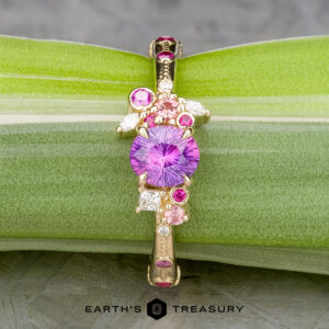 The Linnea ring in 14K yellow gold features a 1.15-carat round Ceylon sapphire centerpiece, accented by pink and white stones, displayed on a green leaf with the Earth's Treasury logo below.