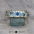 The Thin Borealis Ring in 14k yellow gold, featuring blue and teal gemstones, is displayed upright on a rough green stone. The "Earth's Treasury" logo appears at the bottom.