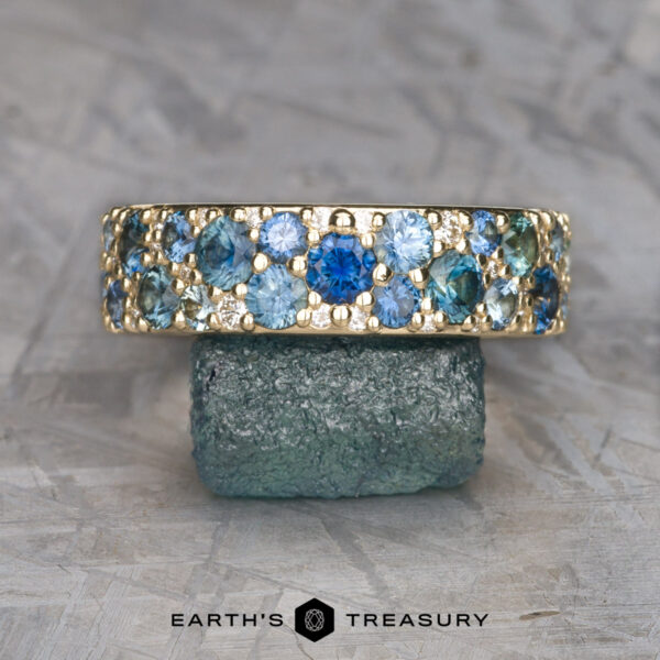 The Thin Borealis Ring in 14k yellow gold The Thin Borealis Ring in 14k yellow gold, featuring blue and teal gemstones, is displayed upright on a rough green stone. The "Earth's Treasury" logo appears at the bottom.