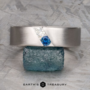 The "Parnis" ring in 14k brushed white gold features a white and a blue gemstone set diagonally, shown atop a rough blue-green crystal.