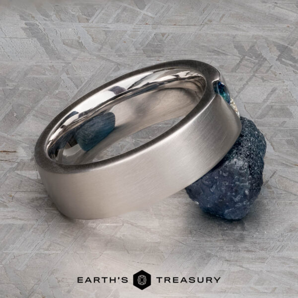 The "Parnis" ring in 14k white gold, brushed The "Parnis" ring in 14k brushed white gold with a blue gemstone is displayed against a rough blue mineral on a textured grey surface. The Earth’s Treasury logo appears at the bottom.