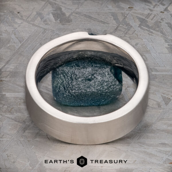 The "Parnis" ring in 14k white gold, brushed A brushed 14k white gold "Parnis" ring encircles a rough blue gemstone, set on a textured gray surface with the text "EARTH'S TREASURY" below.
