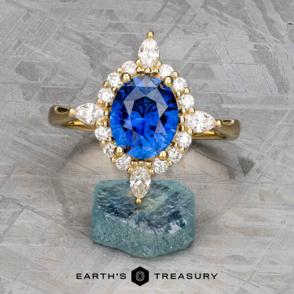The "Francoise" ring in 18k yellow gold with 2.28-carat Ceylon sapphire The "Francoise" ring in 18k yellow gold, featuring a 2.28-carat oval Ceylon sapphire surrounded by clear stones, is displayed on a rough blue mineral with the "Earth's Treasury" logo below.