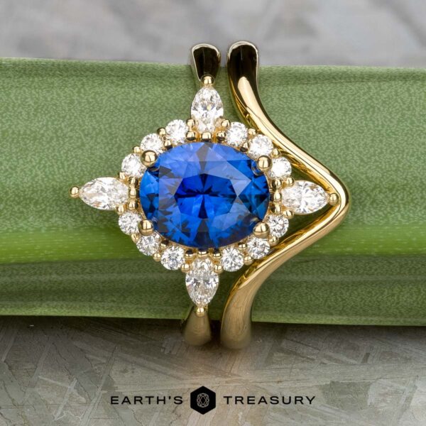 The "Francoise" ring in 18k yellow gold with 2.28-carat Ceylon sapphire, alongside the custom-fit contour band in 18k yellow gold The "Francoise" ring in 18k yellow gold features a 2.28-carat Ceylon sapphire, accented by round and marquise white stones, with its custom-fit contour band, shown on a green background with the "Earth’s Treasury" logo.