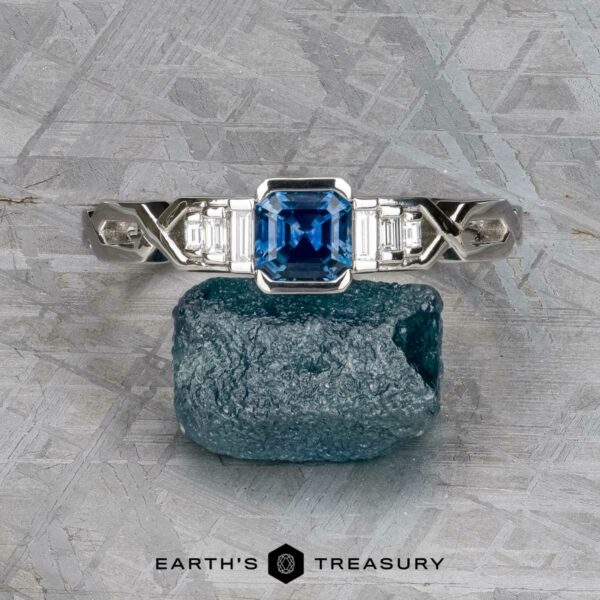 The "Pallas" ring in platinum with 0.73-carat Montana sapphire A platinum "Pallas" ring featuring a 0.73-carat Montana sapphire is displayed upright on a rough blue stone, set against a textured gray background.