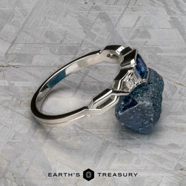The "Pallas" ring in platinum with 0.73-carat Montana sapphire The "Pallas" ring in platinum with a 0.73-carat Montana sapphire stands upright on a rough blue mineral, set on a textured gray surface with the “EARTH'S TREASURY” logo at the bottom.
