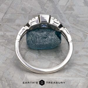 A platinum "Pallas" ring set with a 0.73-carat Montana sapphire stands upright against a rough blue stone on a textured gray surface, with the text "Earth's Treasury" below.