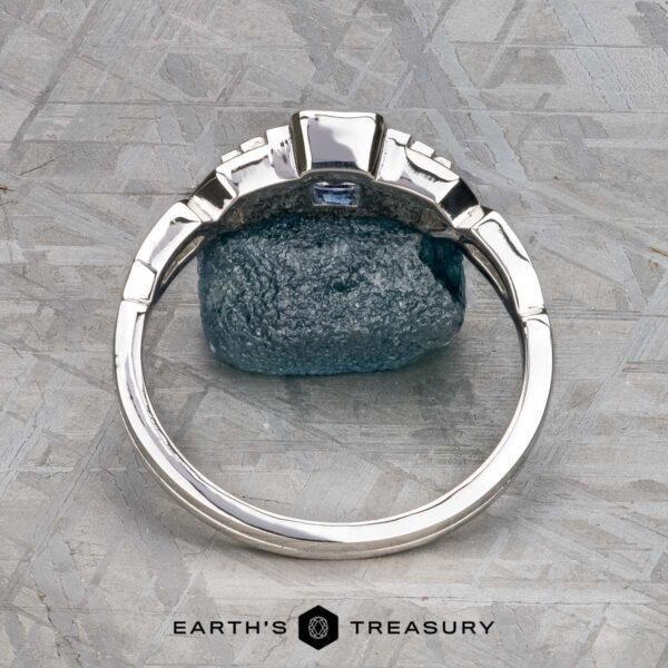 The "Pallas" ring in platinum with 0.73-carat Montana sapphire A platinum "Pallas" ring set with a 0.73-carat Montana sapphire stands upright against a rough blue stone on a textured gray surface, with the text "Earth's Treasury" below.