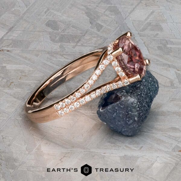 The "Senna" ring in 14k rose gold with 2.20-carat Umba sapphire The "Senna" ring in 14k rose gold, featuring a 2.20-carat pink Umba sapphire and diamond accents, is displayed leaning on a rough gray stone with the Earth's Treasury logo at the bottom.