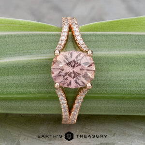 The "Senna" ring features a 2.20-carat round pink Umba sapphire set in 14k rose gold with a split diamond band, shown on a green striped leaf.