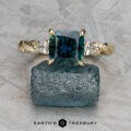 The "Lenore" ring in 14k yellow gold, featuring a 2.51-carat Australian sapphire and two side stones, is displayed on rough blue mineral with a geometric background. Earth's Treasury logo appears at the bottom.