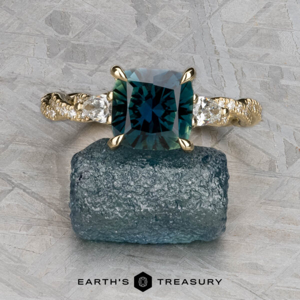 The "Lenore" ring in 14k yellow gold with 2.51-carat Australian sapphire The "Lenore" ring in 14k yellow gold, featuring a 2.51-carat Australian sapphire and two side stones, is displayed on rough blue mineral with a geometric background. Earth's Treasury logo appears at the bottom.