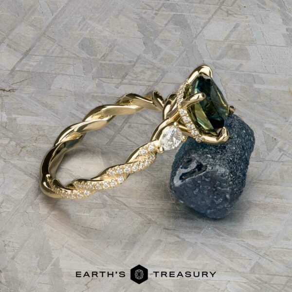 The "Lenore" ring in 14k yellow gold with 2.51-carat Australian sapphire The "Lenore" ring in 14k yellow gold, featuring a 2.51-carat Australian sapphire and diamond accents, rests on dark rough stone. A textured metallic backdrop highlights the piece, with the "Earth's Treasury" logo at the bottom.