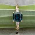 The "Lenore" ring in 14k yellow gold features a 2.51-carat Australian sapphire and two pear-shaped side stones, shown on a green leaf background.
