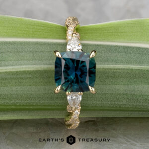The "Lenore" ring in 14k yellow gold features a 2.51-carat Australian sapphire and two pear-shaped side stones, shown on a green leaf background.