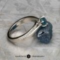 The "Astrid" ring in platinum with a 0.98-carat Montana sapphire rests beside a rough blue stone on a light textured surface; the "Earth's Treasury" logo appears at the bottom.
