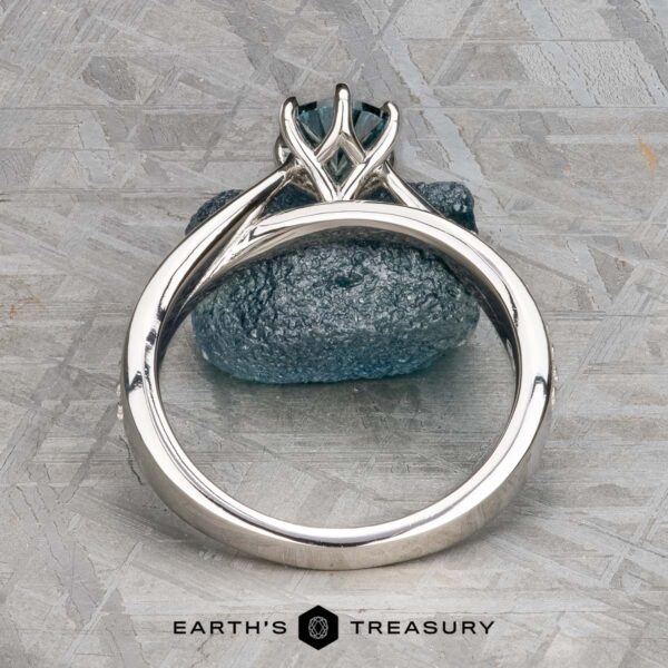 The "Astrid" ring in platinum with 0.98-carat Montana sapphire The "Astrid" ring in platinum features a 0.98-carat Montana sapphire set upright with prongs, photographed on a textured gray surface beside a blue mineral and the "Earth's Treasury" logo at the bottom.