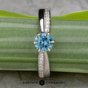 The "Astrid" ring in platinum features a 0.98-carat Montana sapphire and small clear accent stones, displayed on a green leaf with "Earth's Treasury" branding below.