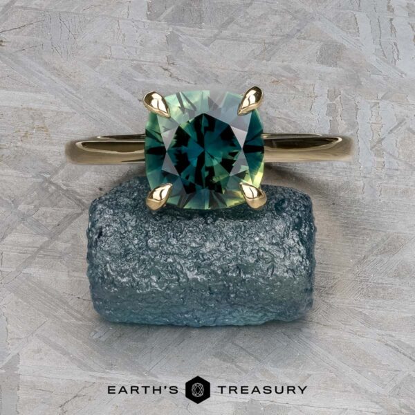 The Cathedral "Begonia" ring in 14k yellow gold with 2.27-carat Australian sapphire The Cathedral "Begonia" ring in 14k yellow gold with a 2.27-carat Australian sapphire is displayed on a rough blue-green mineral, with the "EARTH'S TREASURY" logo at the bottom.