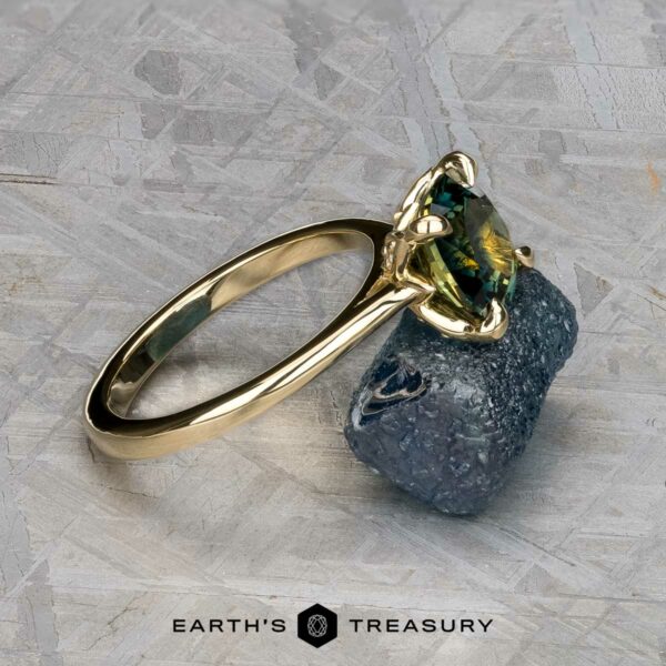 The Cathedral "Begonia" ring in 14k yellow gold with 2.27-carat Australian sapphire The Cathedral "Begonia" ring in 14k yellow gold, featuring a 2.27-carat Australian sapphire, stands upright on a rough blue stone with a textured gray background and the text "Earth's Treasury" below.