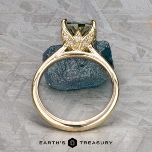 The Cathedral "Begonia" ring in 14k yellow gold, featuring a 2.27-carat Australian sapphire, is displayed upright before a rough stone. The "EARTH'S TREASURY" logo appears at the bottom.