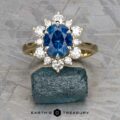 The "Angelica" ring, crafted in 14k white and yellow gold with a 2.30-carat Montana sapphire surrounded by diamonds, is displayed on a rough blue stone. The Earth's Treasury logo appears below.
